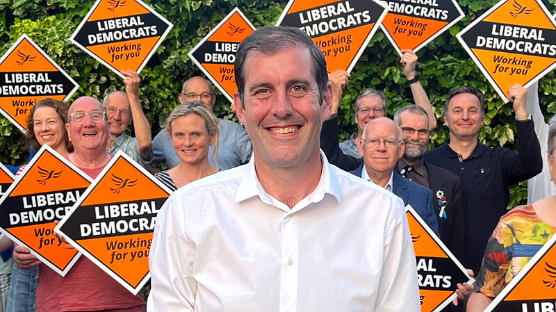 Lee Dillon standing in front of a group of Liberal Democrat activists holding orange diamond signs with the words "Liberal Democrats" on them