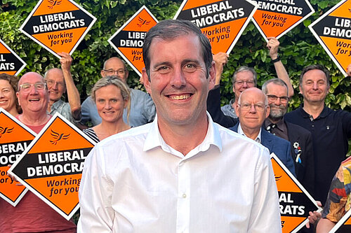 Lee Dillon standing in front of a group of Liberal Democrat activists holding orange diamond signs with the words "Liberal Democrats" on them